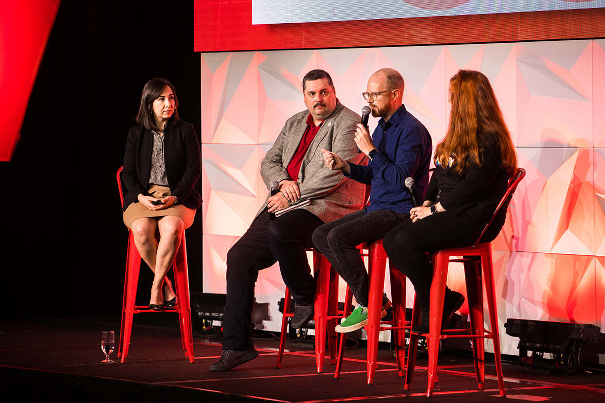A photo of four panelists sitting on the stage on red stools during the panel.